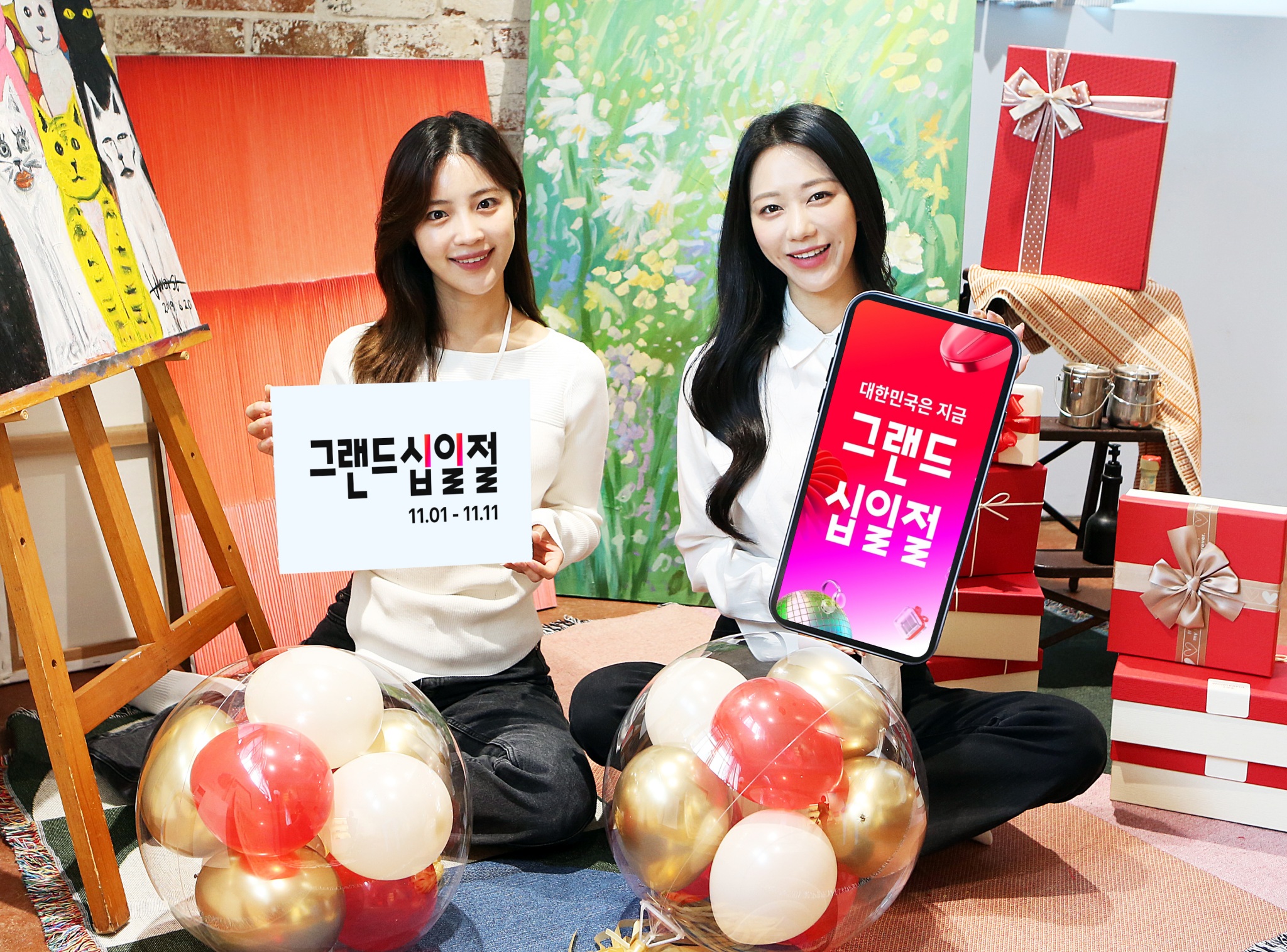 여자 모델들이 선물 상자 가득한 곳에서 11월1일부터 11일까지 진행되는 그랜드십일절 행사 안내 판넬을 들고 있다